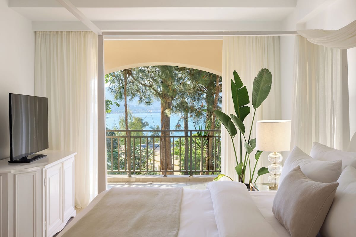 Quarto de hotel com uma cama grande, uma televisão em um móvel branco, uma varanda com vista para o mar e um vaso de planta.