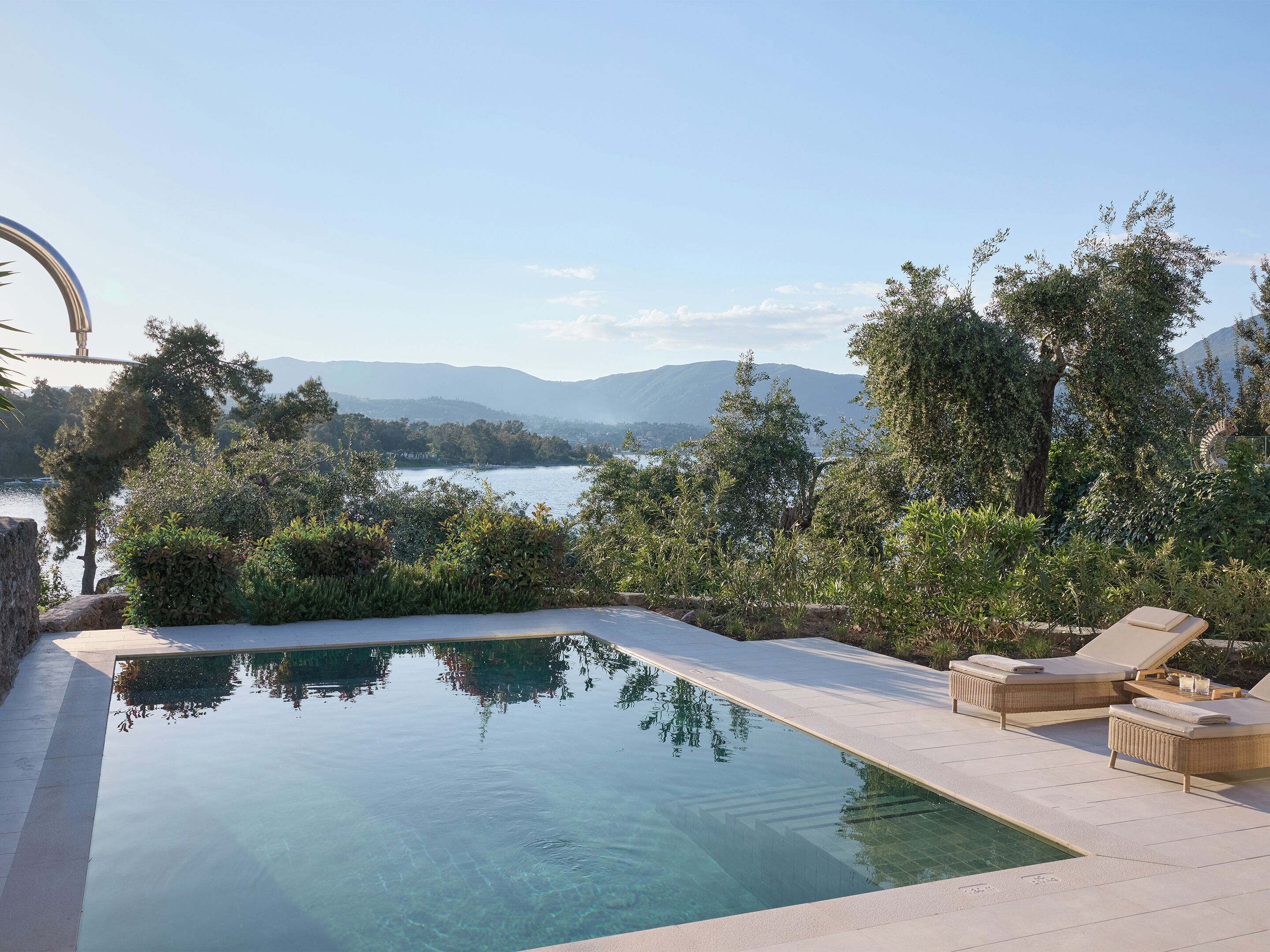 A clear swimming pool with lounge chairs and a view of a lake and mountains.