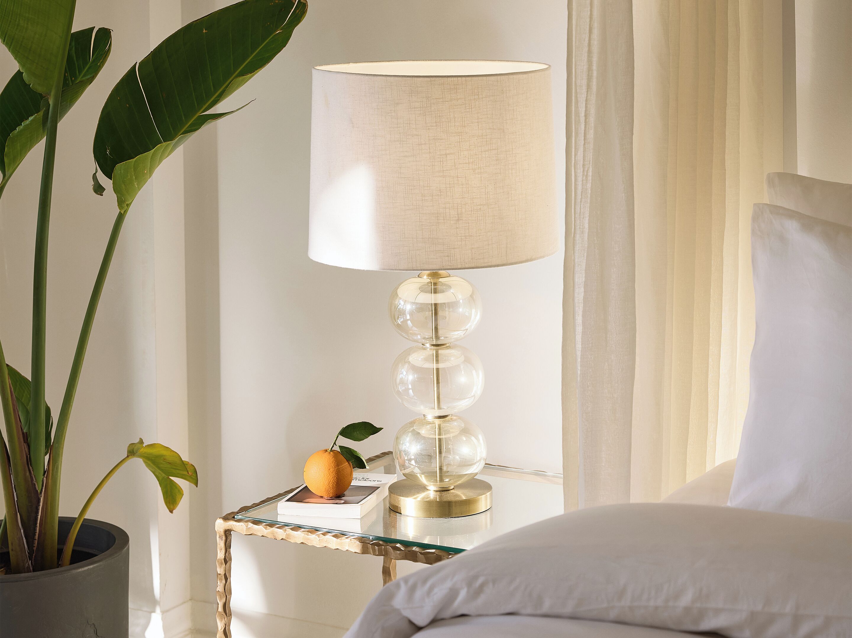 Habitación de hotel ordenada, con una cama, una mesita de noche con lámpara, una planta en maceta y un detalle decorativo.