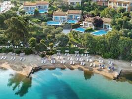 Una vista aerea di un resort con diverse ville, una spiaggia attrezzata con lettini e ombrelloni, e una grande piscina.
