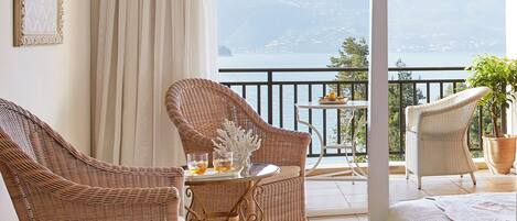 Un balcón con mobiliario de mimbre, una mesa con bebidas y vistas a un lago y montañas.
