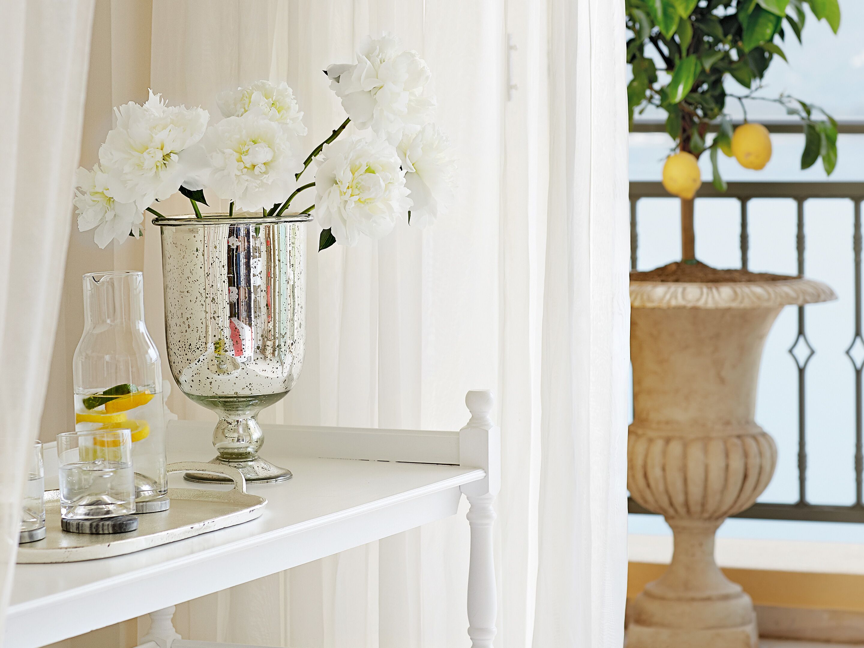 A white table with a decorative silver vase containing white flowers, a glass bottle with a lemon slice, and a glass of water.