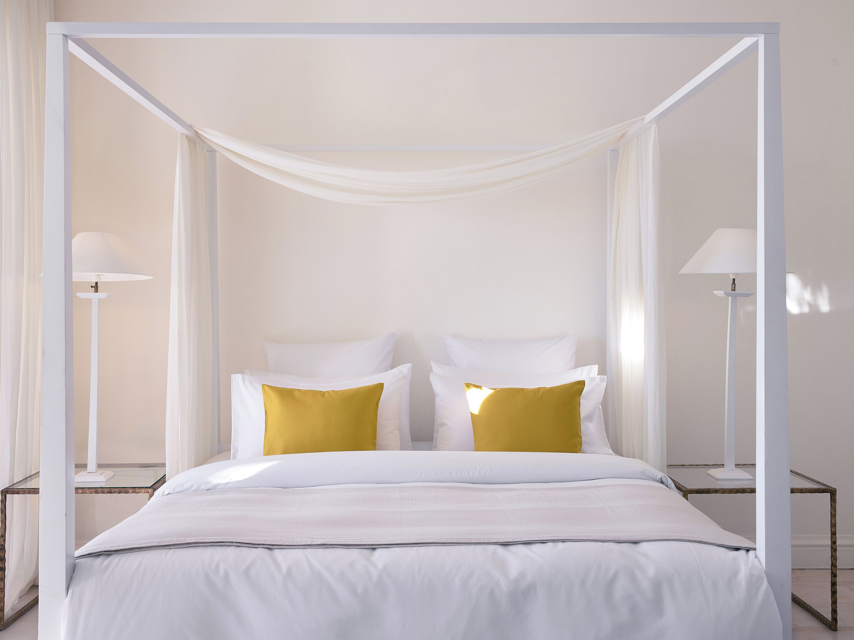 Ein Himmelsbett mit weißer Bettwäsche und zwei gelben Kissen, flankiert von zwei Nachttischen mit Lampen.