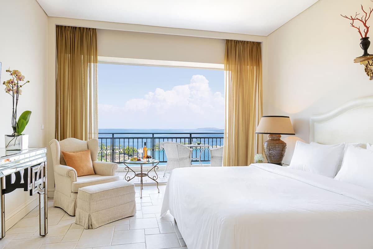 Quarto de hotel com uma cama grande, um espaço de estar com uma poltrona e apoio para os pés, uma pequena mesa com um vaso de flores e vista para o mar através de uma varanda.