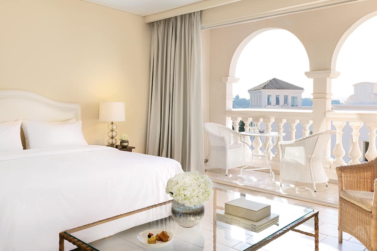 Quarto de hotel com uma cama grande, uma mesa de centro de vidro com um livro e um prato de comida, cadeiras de vime branco e vista para um prédio através de um arco.