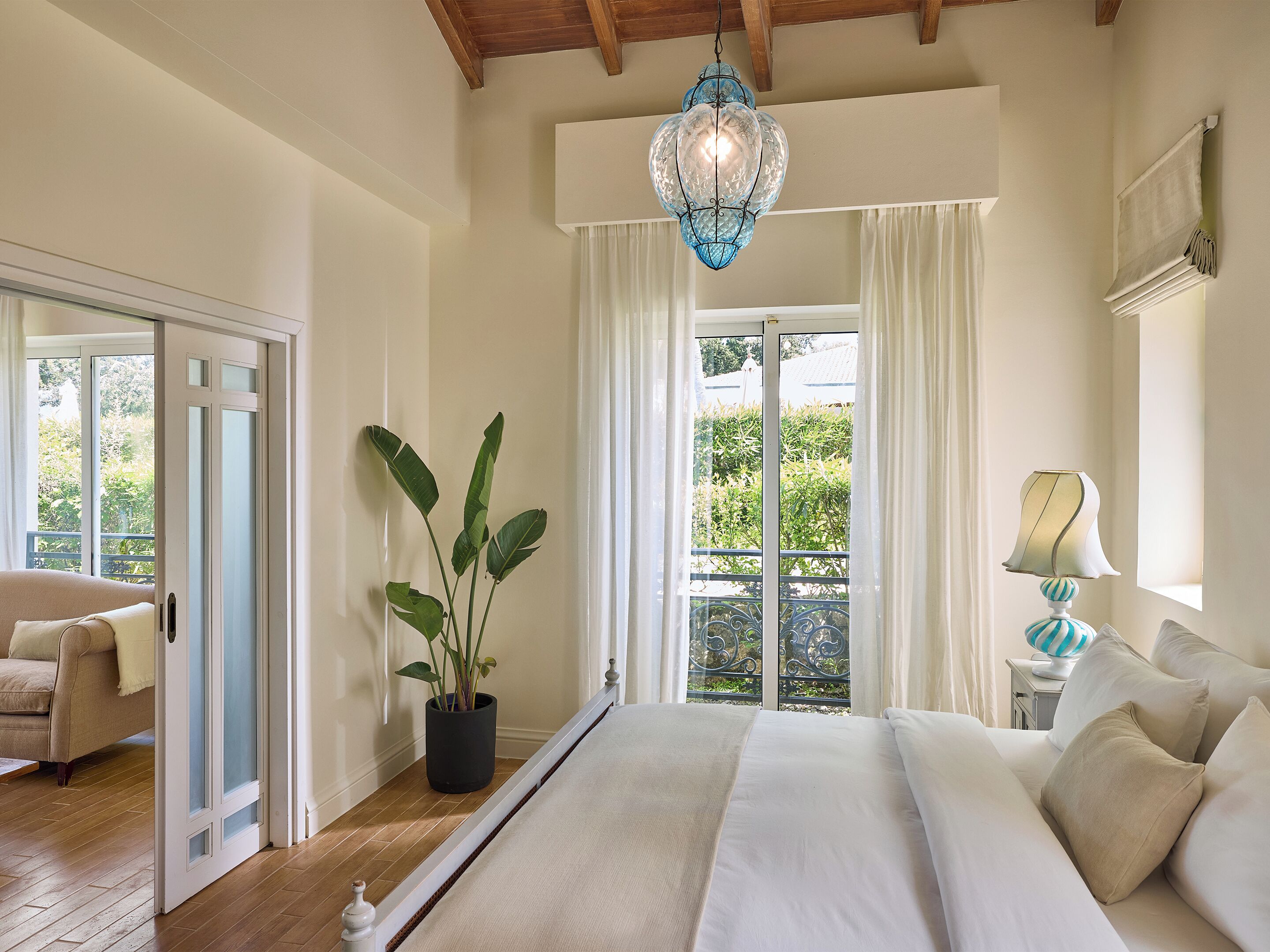 A bedroom with a large bed, a potted plant, a hanging light fixture, and a view of greenery outside.