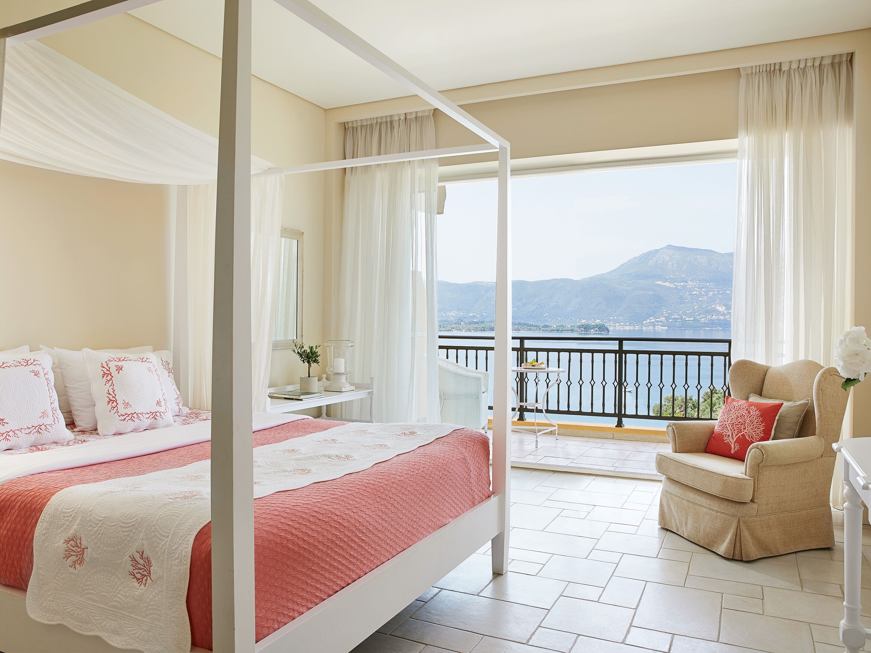 Un lit à baldaquin avec une couvre-lit rouge et blanche, une petite table ornée d’un vase et d’une bougie, une chaise avec un coussin rouge, et un balcon offrant une vue sur la mer et les montagnes.