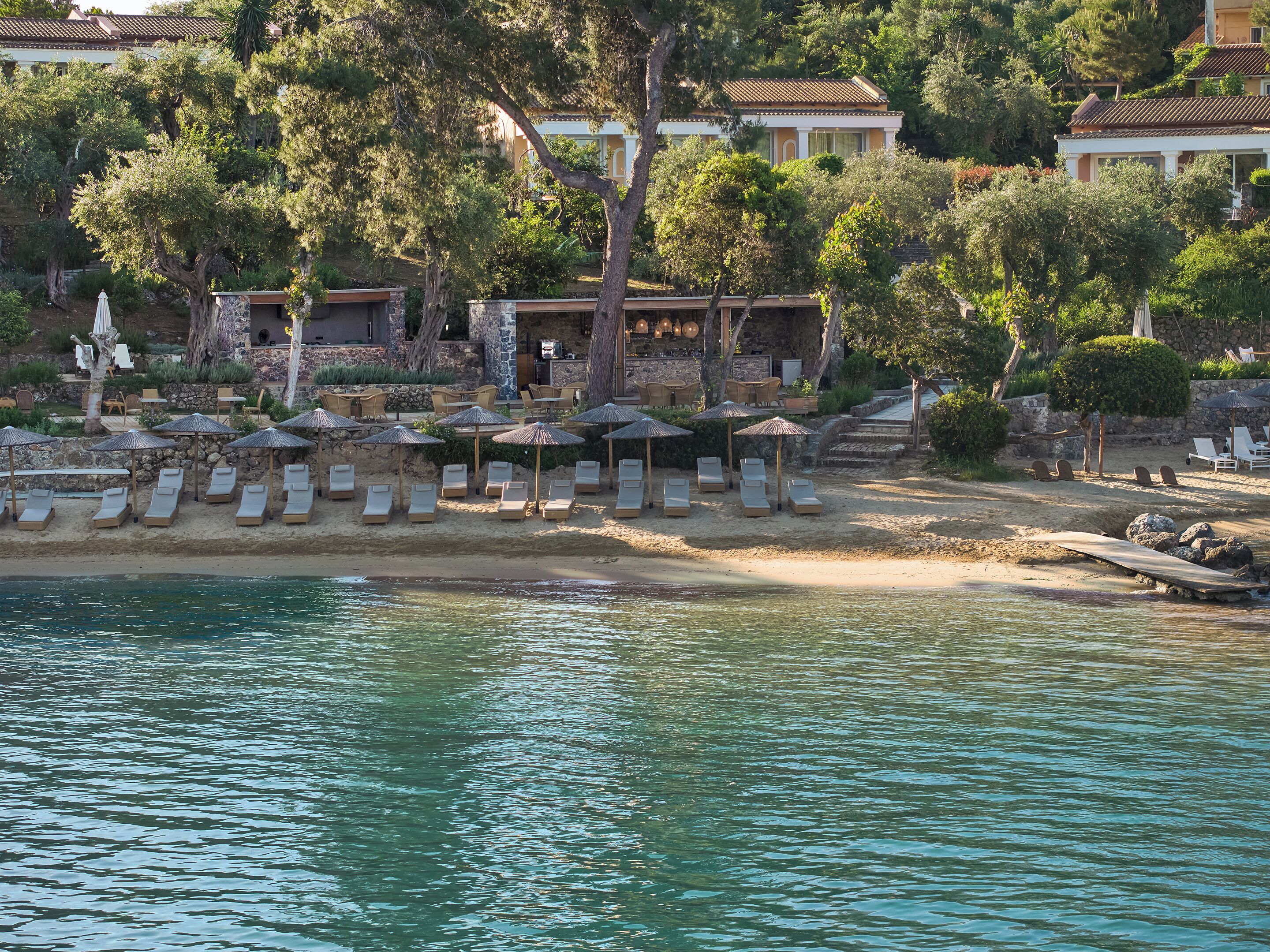 A coastal resort with sun loungers and umbrellas on the beach, a pool, and a stone building with a tiled roof.