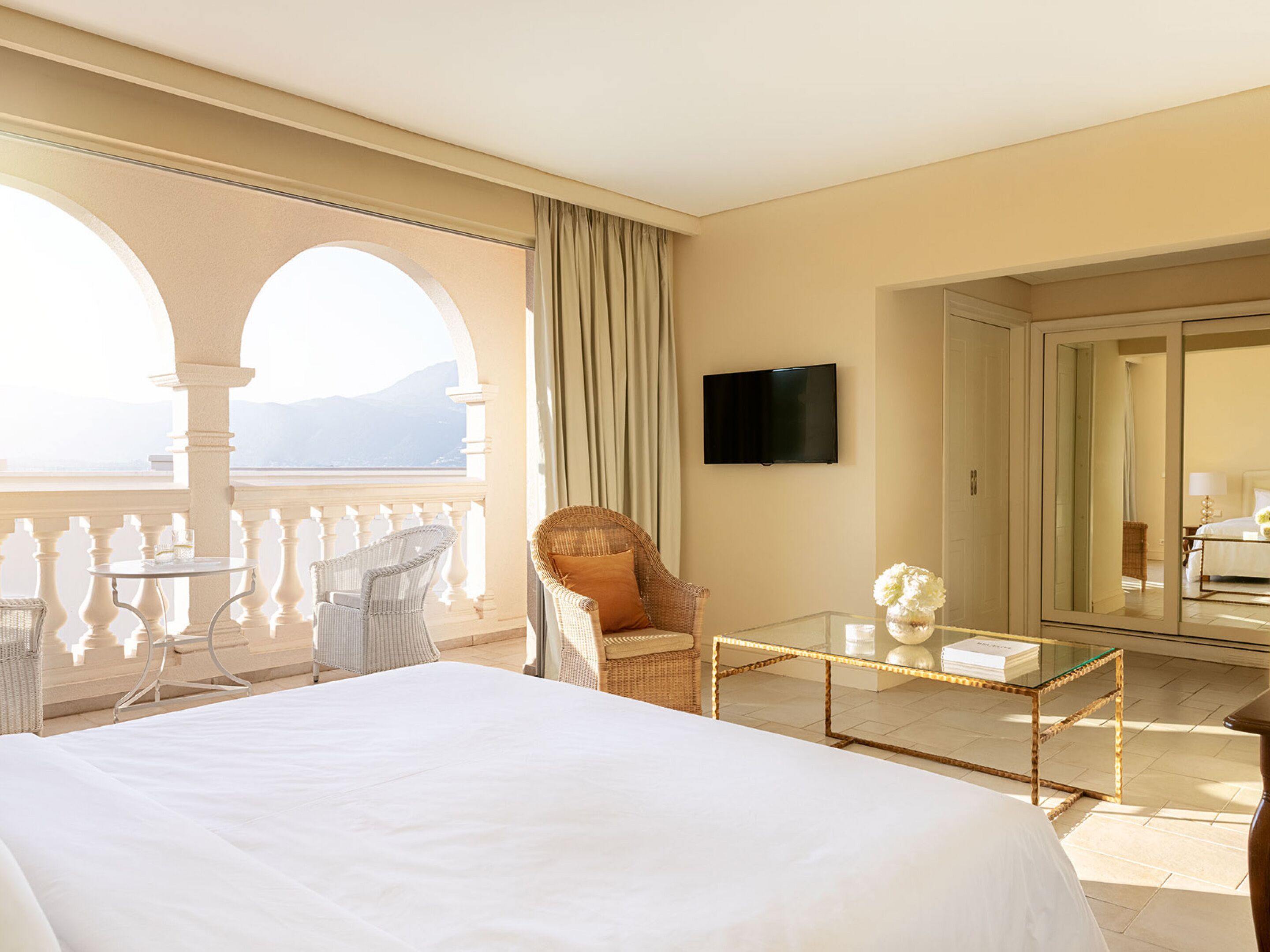 Habitación de hotel con una cama grande, un balcón con mobiliario de mimbre, televisión y vistas a las montañas.