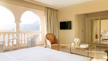 Habitación de hotel con una cama grande, un balcón con mobiliario de mimbre, televisión y vistas a las montañas.