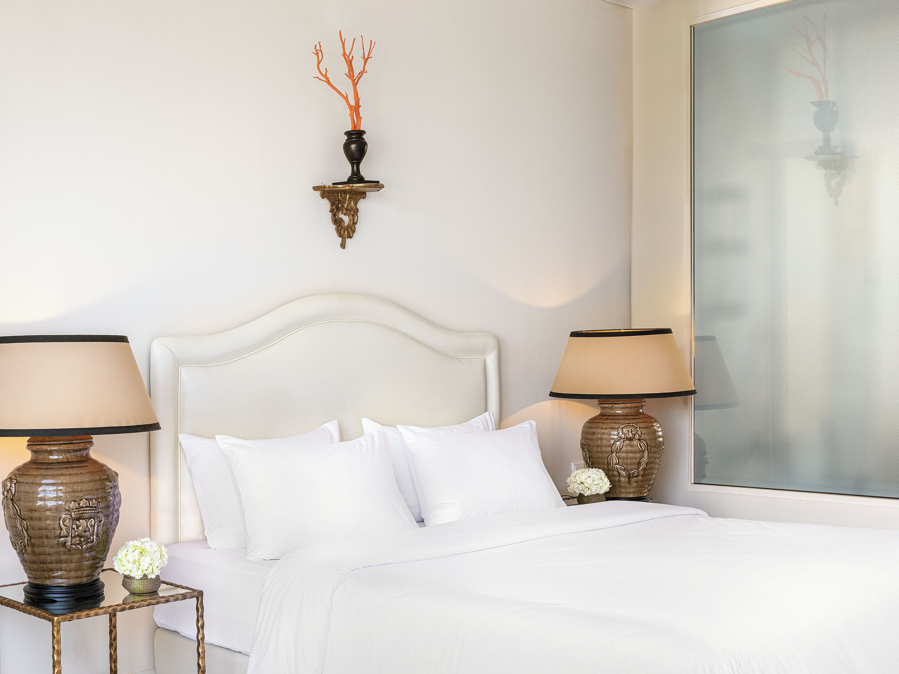 A neatly made bed with white linens, two bedside tables with lamps, a wall-mounted light fixture, and a large window with a view.