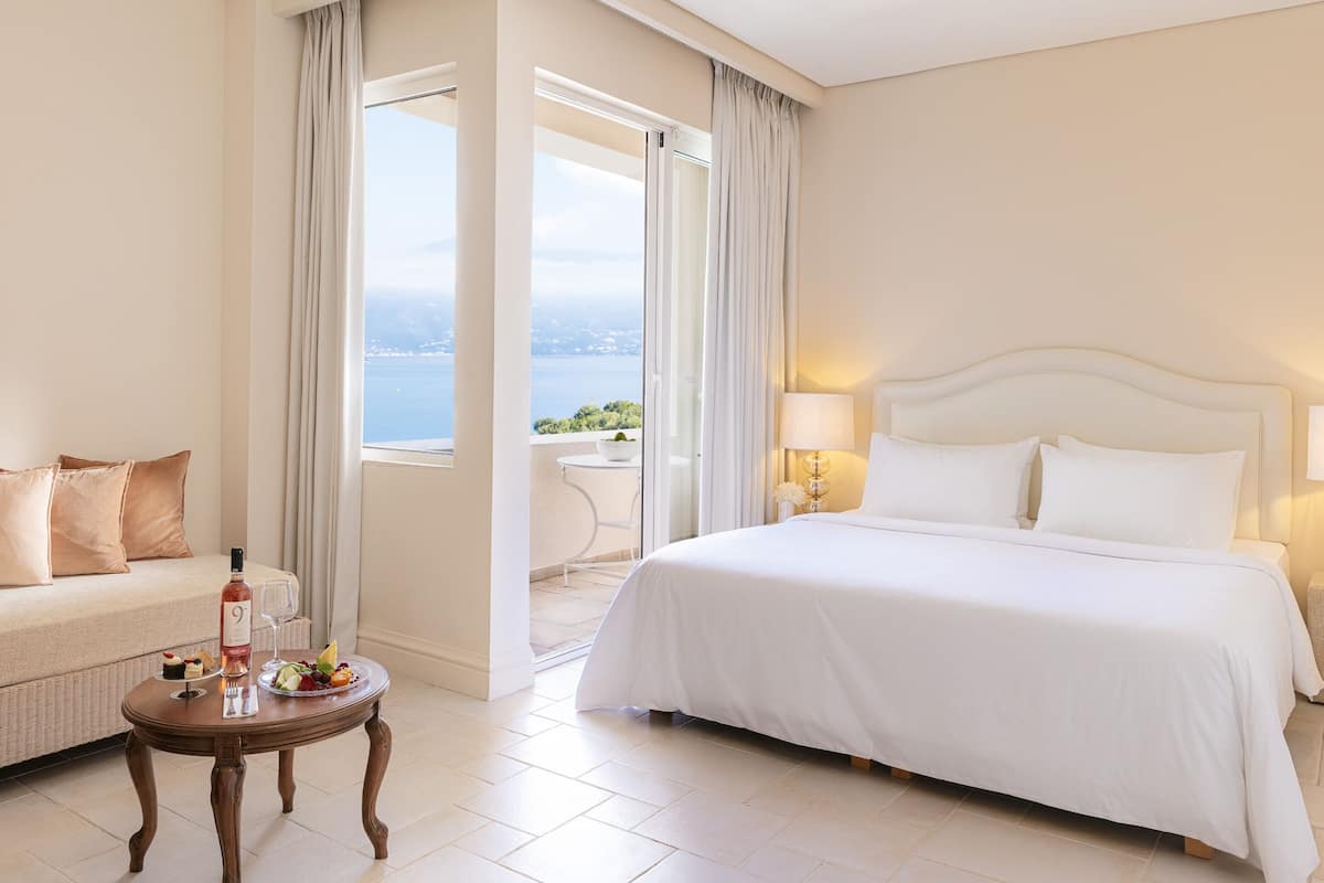 Quarto de hotel com uma cama grande, um sofá, uma mesa com garrafa e copos, e vista para o mar.