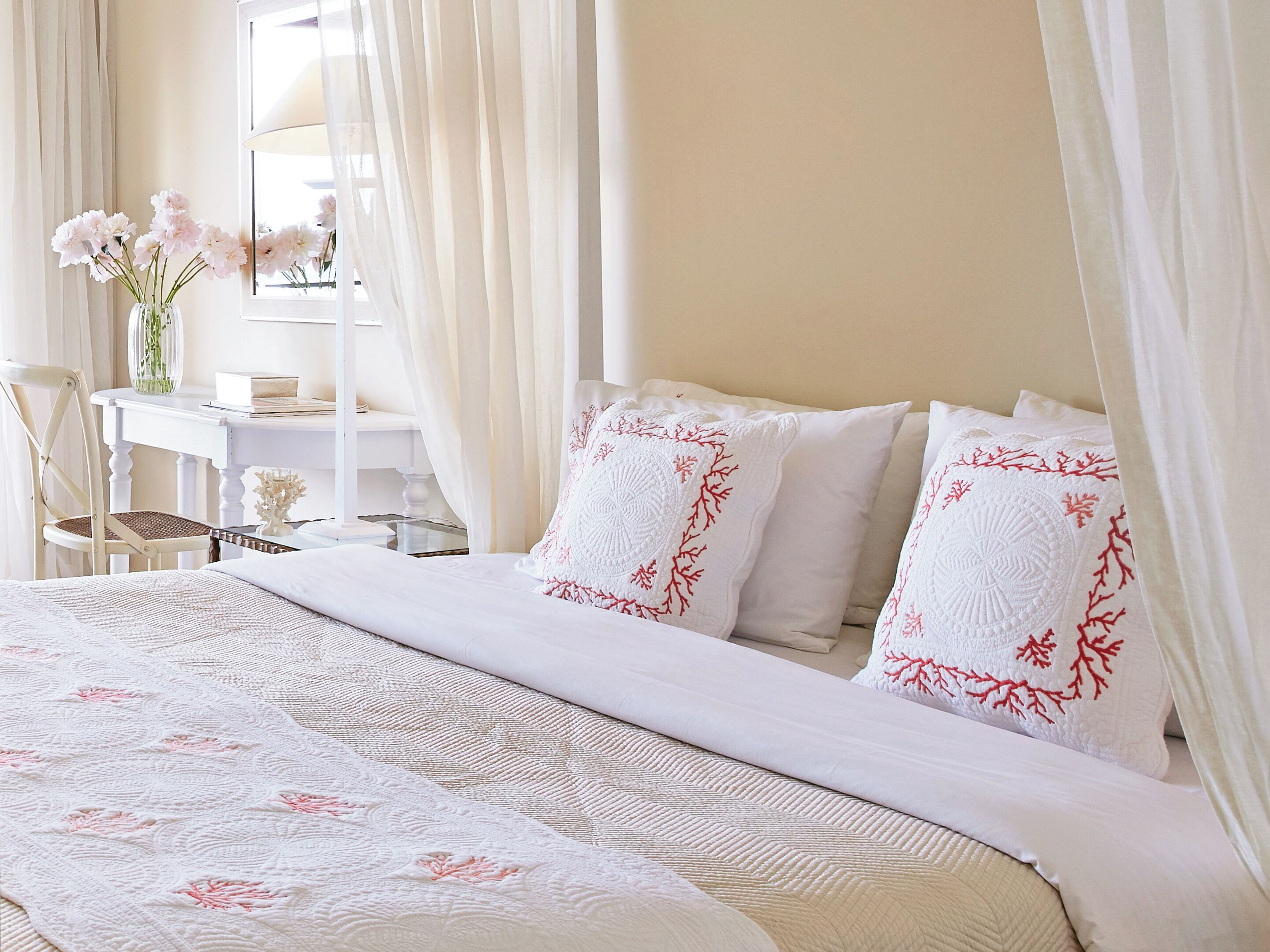 A neatly made bed with white linens and red floral pillows, a white desk with a vase of flowers, and sheer curtains.