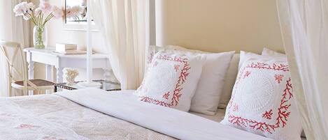 Una cama bien hecha con sábanas blancas y almohadas rojas con estampado floral, un escritorio blanco con un florero y cortinas de encaje.