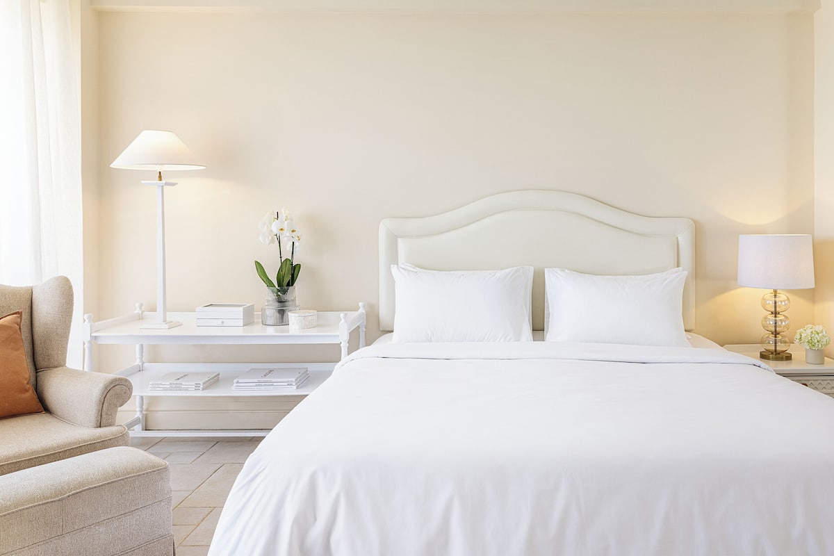 Quarto de hotel com uma cama grande, uma mesinha de cabeceira com abajur, uma pequena mesa com um vaso de flores e um sofá.