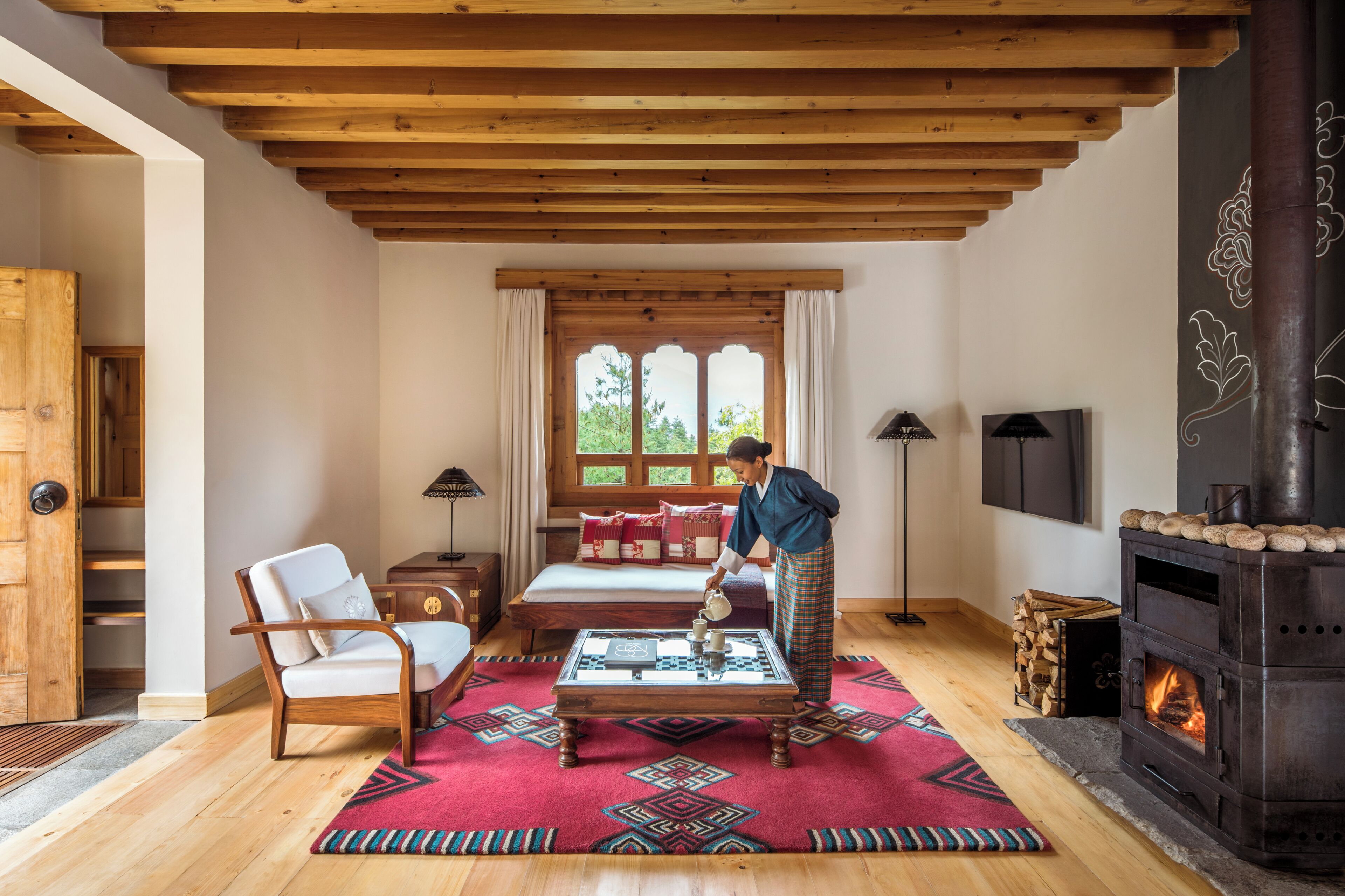 A cozy room with a wooden ceiling, a bed with red and white bedding, a table with a teapot, a chair, and a fireplace.