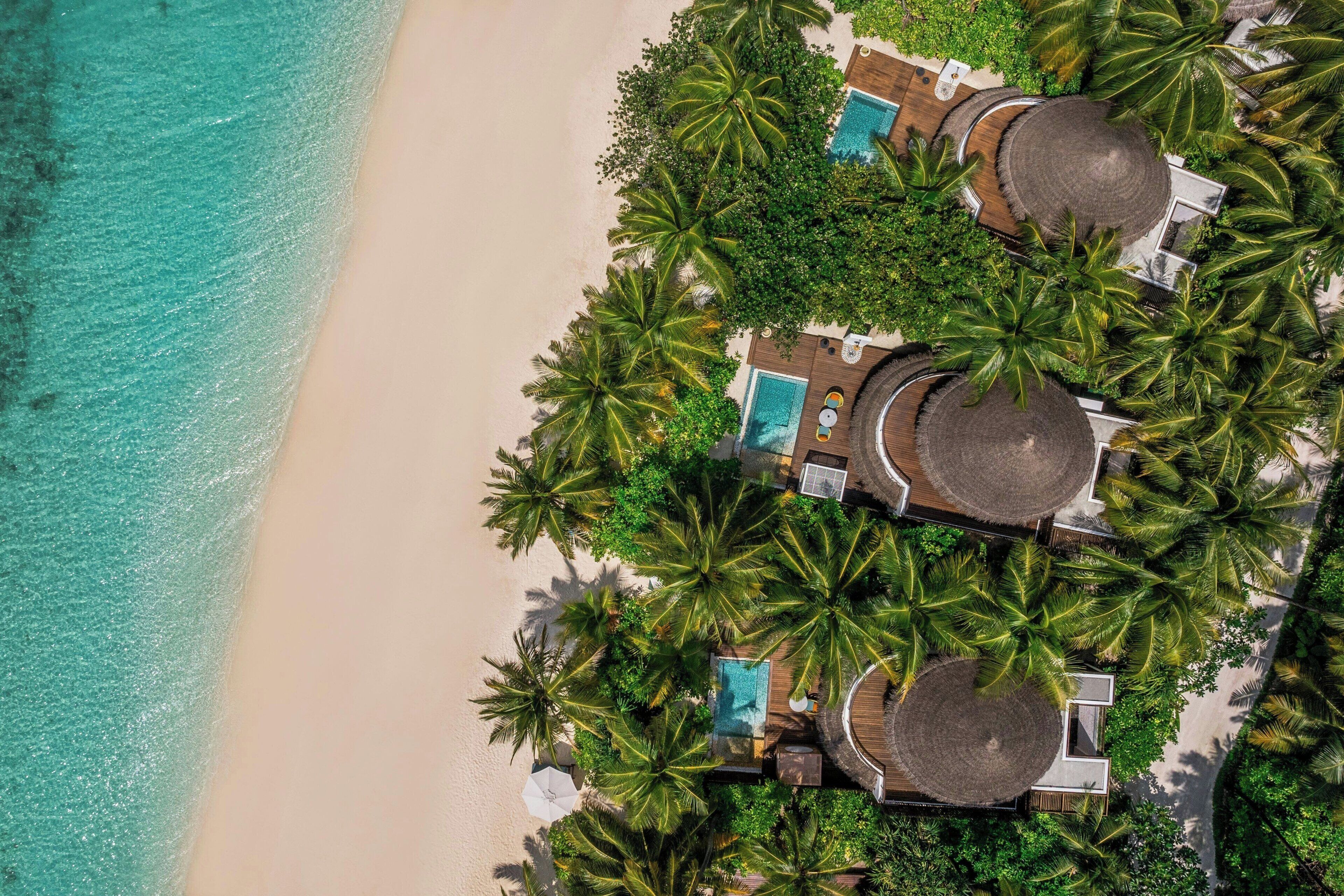 Vista aérea de un resort frente a la playa con varias villas tipo cabaña, cada una con piscina privada y rodeadas de exuberantes palmeras.