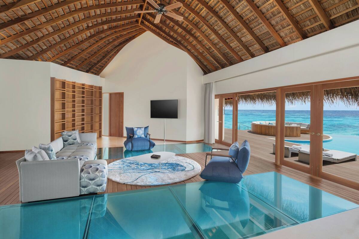 Amplia sala de estar con una mesa de centro de vidrio, asientos con cojines, un televisor de pantalla plana y vistas al océano a través de grandes puertas de cristal.
