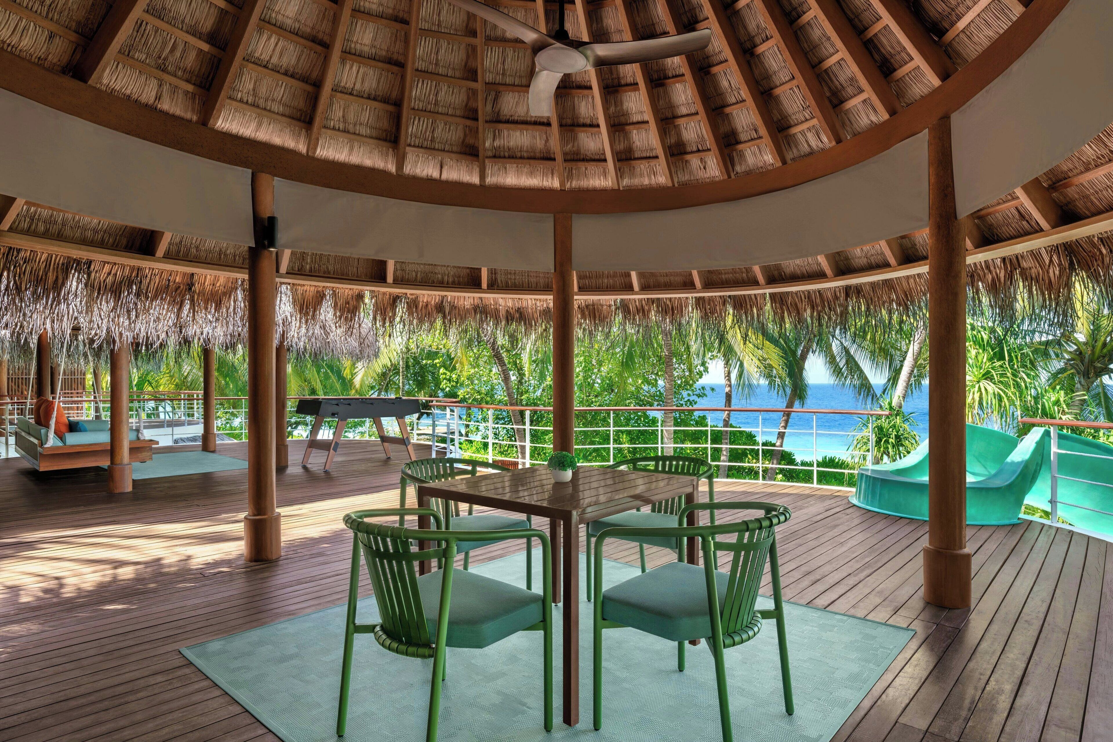 A covered outdoor area with a wooden deck, green chairs, a table, and a thatched roof.