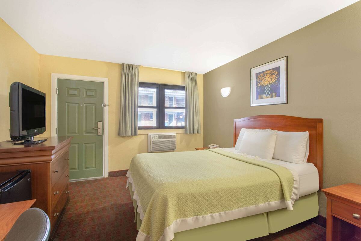 A hotel room with a bed, a television on a dresser, a window with curtains, and a painting on the wall.