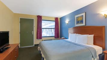 A hotel room with a bed, a television on a wooden dresser, a window with curtains, and a wall-mounted light fixture.
