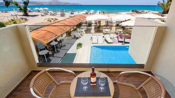 Balcón con sillas de mimbre y una mesa, con vista a una piscina y playa con sombrillas y sillas de descanso.