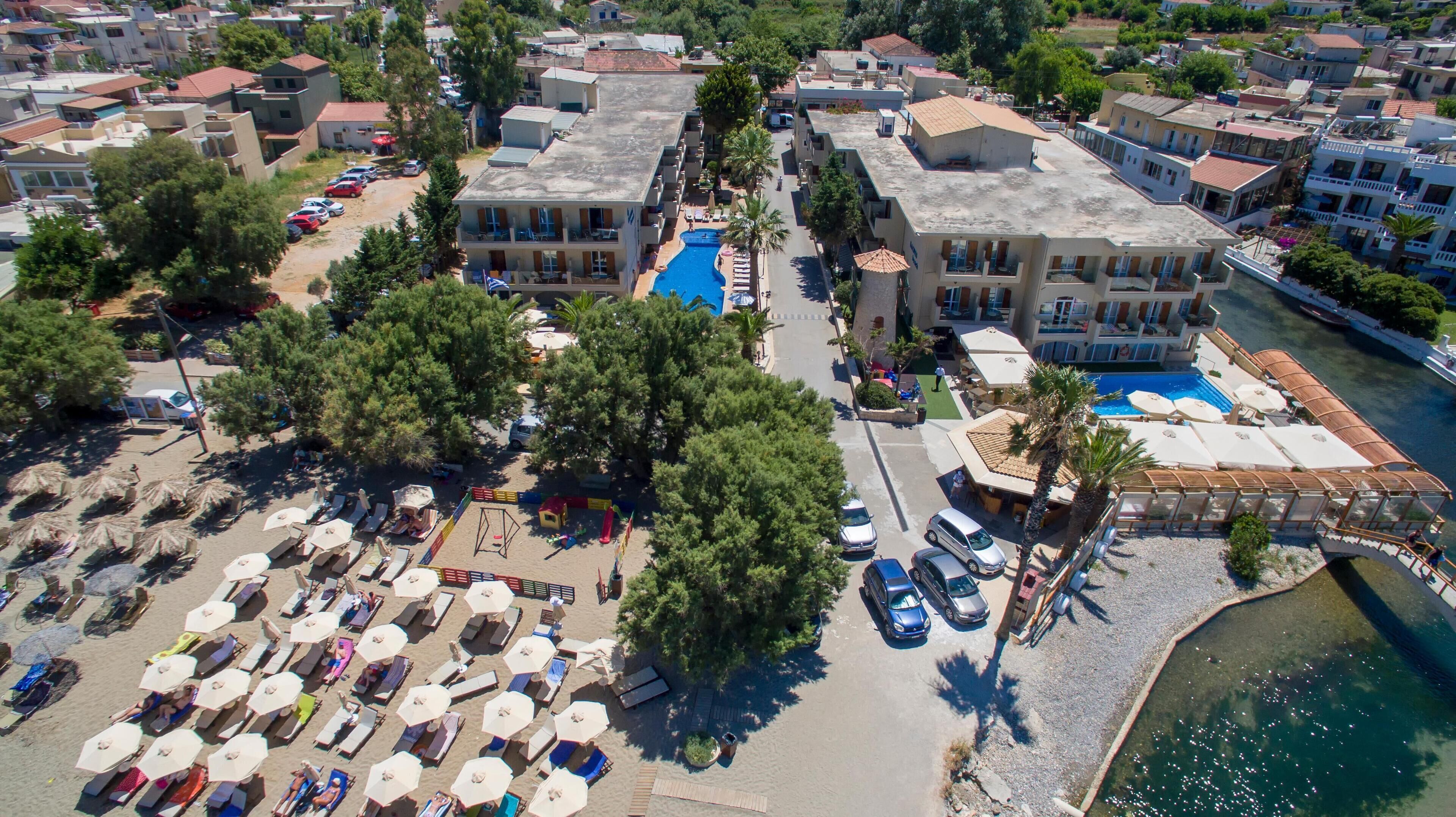 An aerial view of a coastal resort with multiple buildings, a central swimming pool, and a beach area with sun loungers and umbrellas.