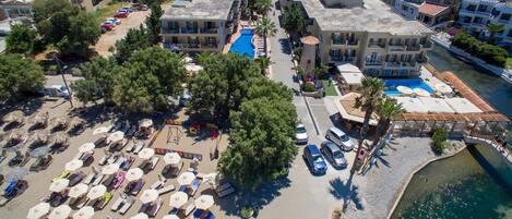 An aerial view of a coastal resort with multiple buildings, a central swimming pool, and a beach area with sun loungers and umbrellas.