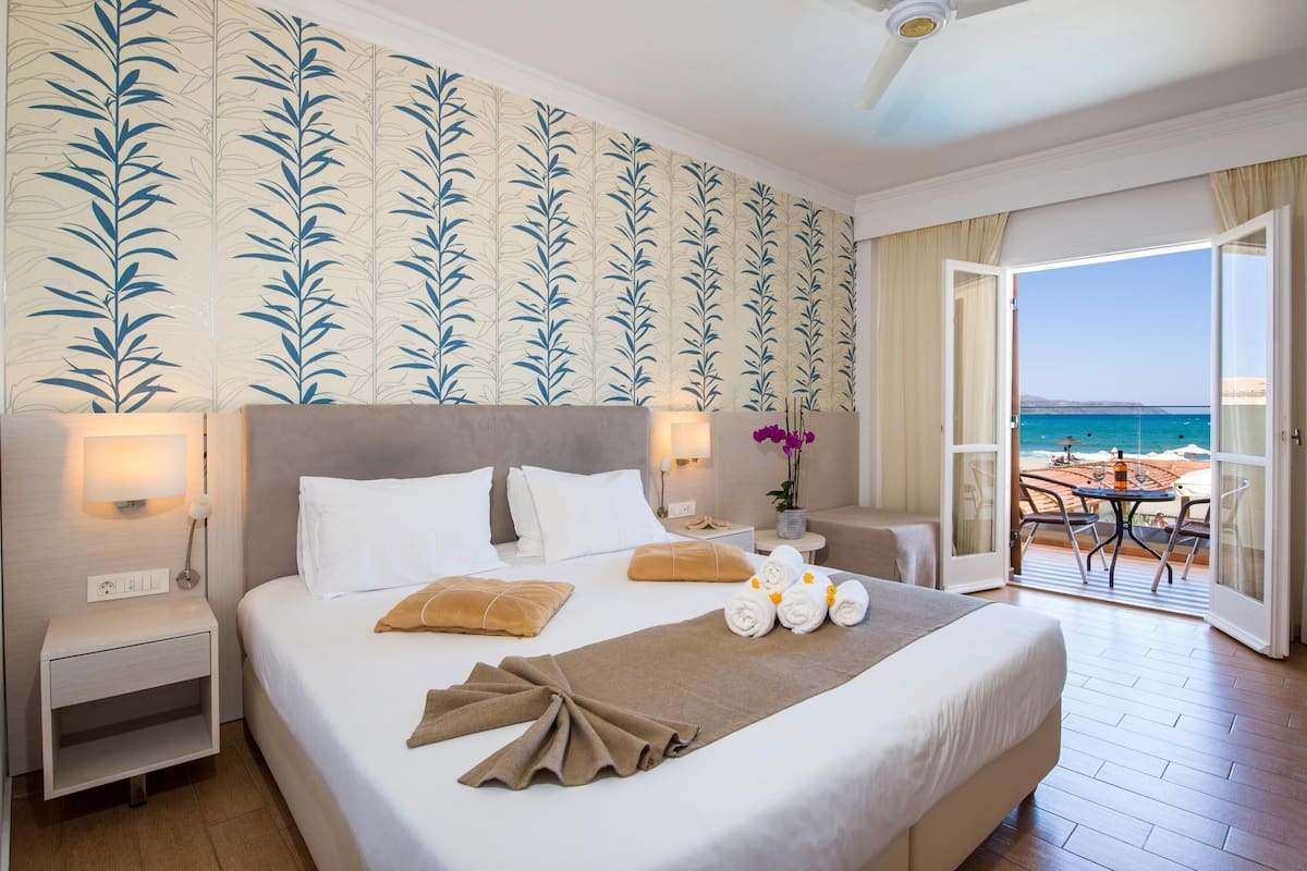 A hotel room with a bed, a nightstand, a lamp, a window with a view of the sea, and a balcony with a table and chairs.