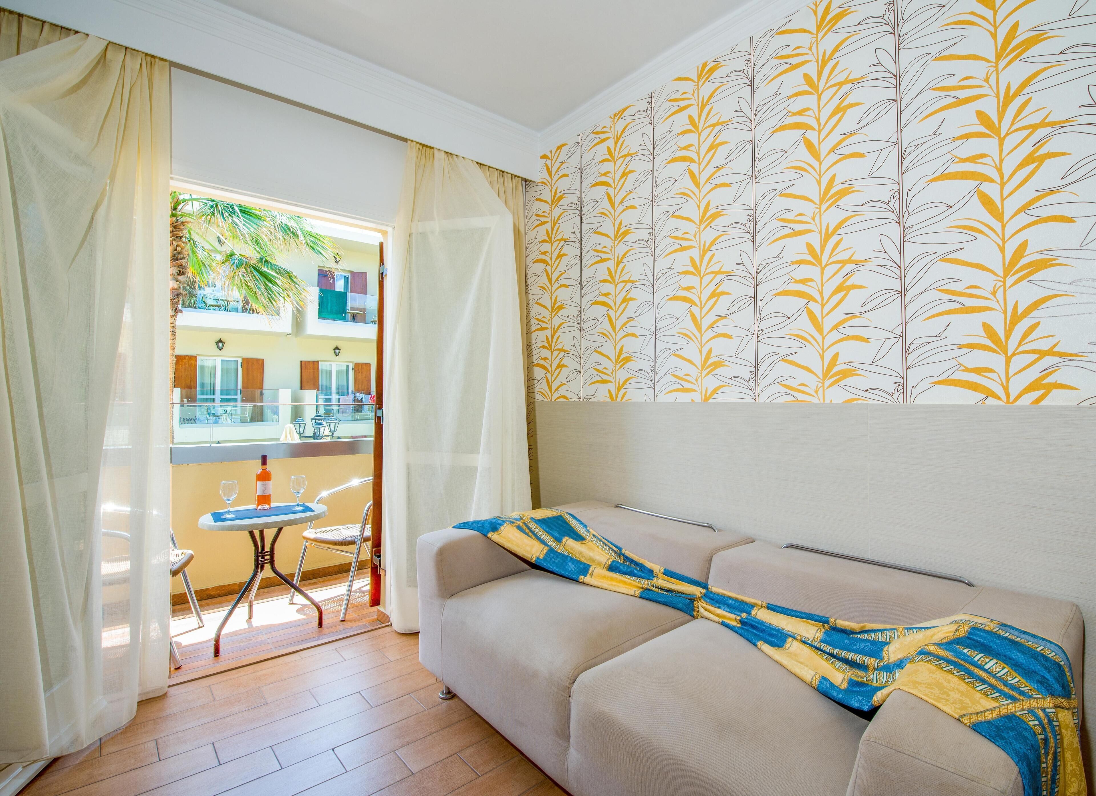 Una habitación con un sofá color beige, una pequeña mesa con superficie de vidrio y una silla. Hay un balcón con vistas a edificios y palmeras.