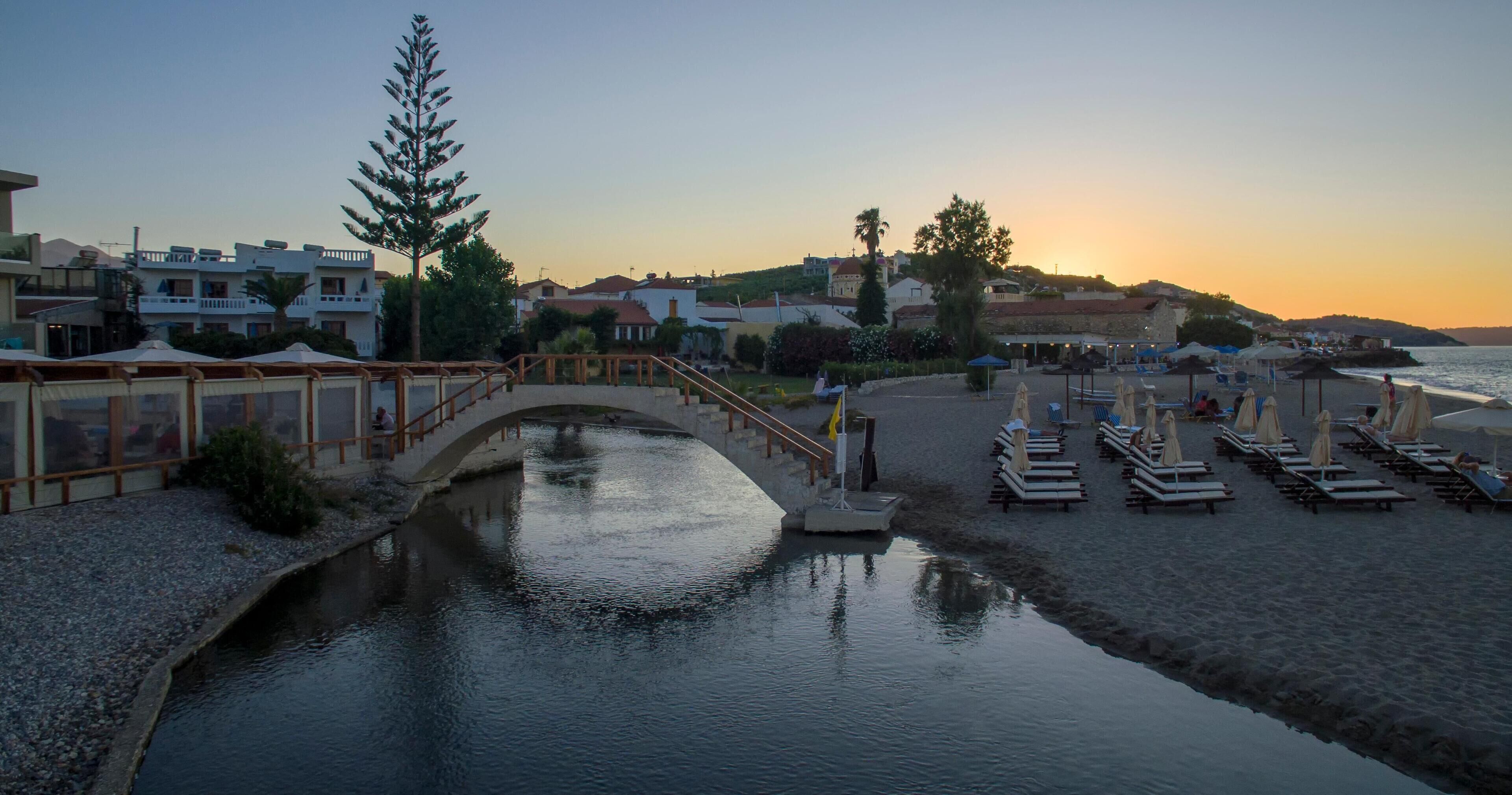 Un hotel a orillas del río, con un puente, sombrillas de playa y sillones de descanso.