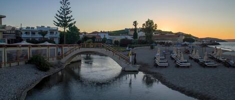 Un hotel a orillas del río, con un puente, sombrillas de playa y sillones de descanso.