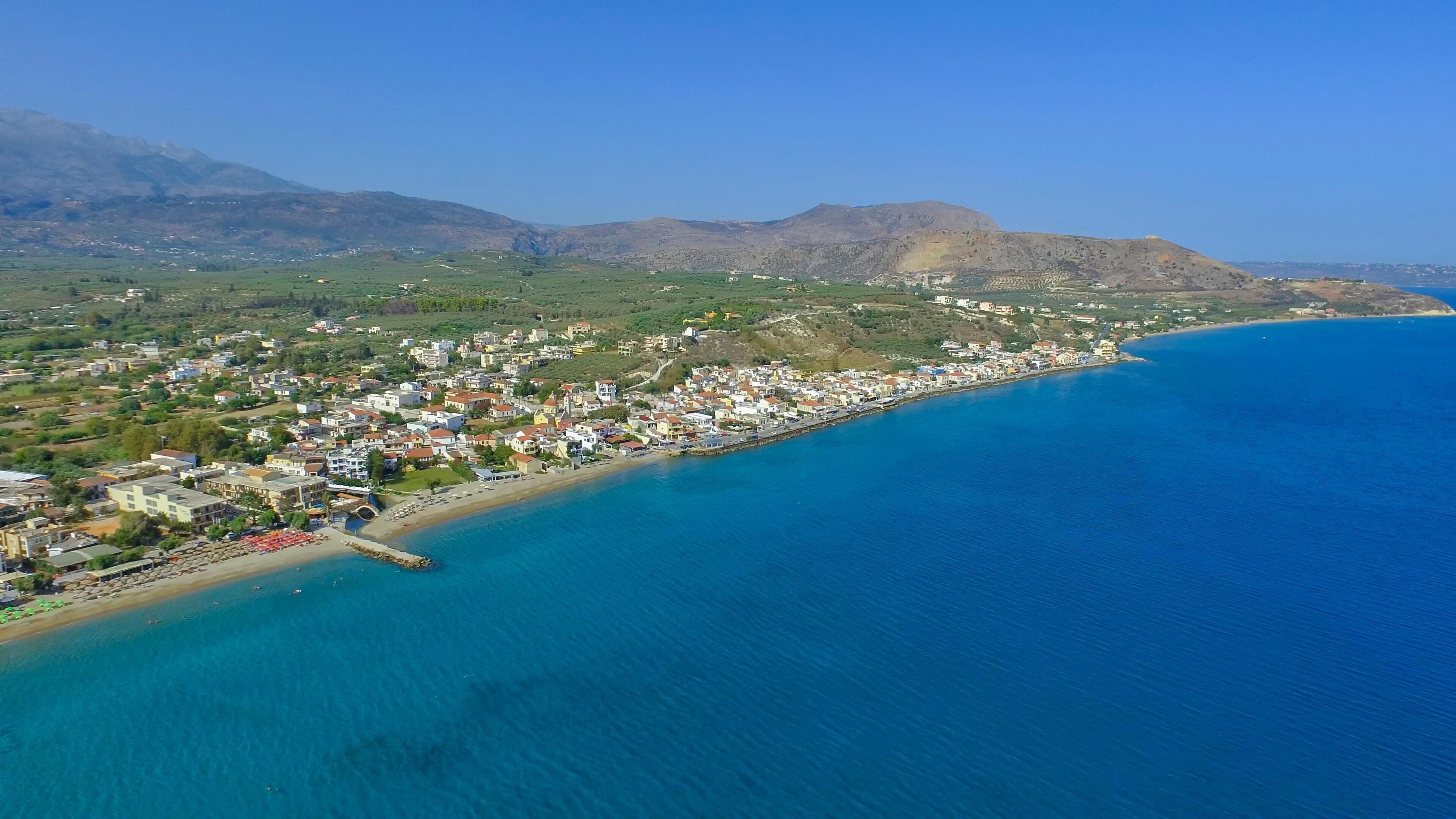 An aerial view of a coastal town with a long beachfront, dense residential area, and mountainous backdrop.