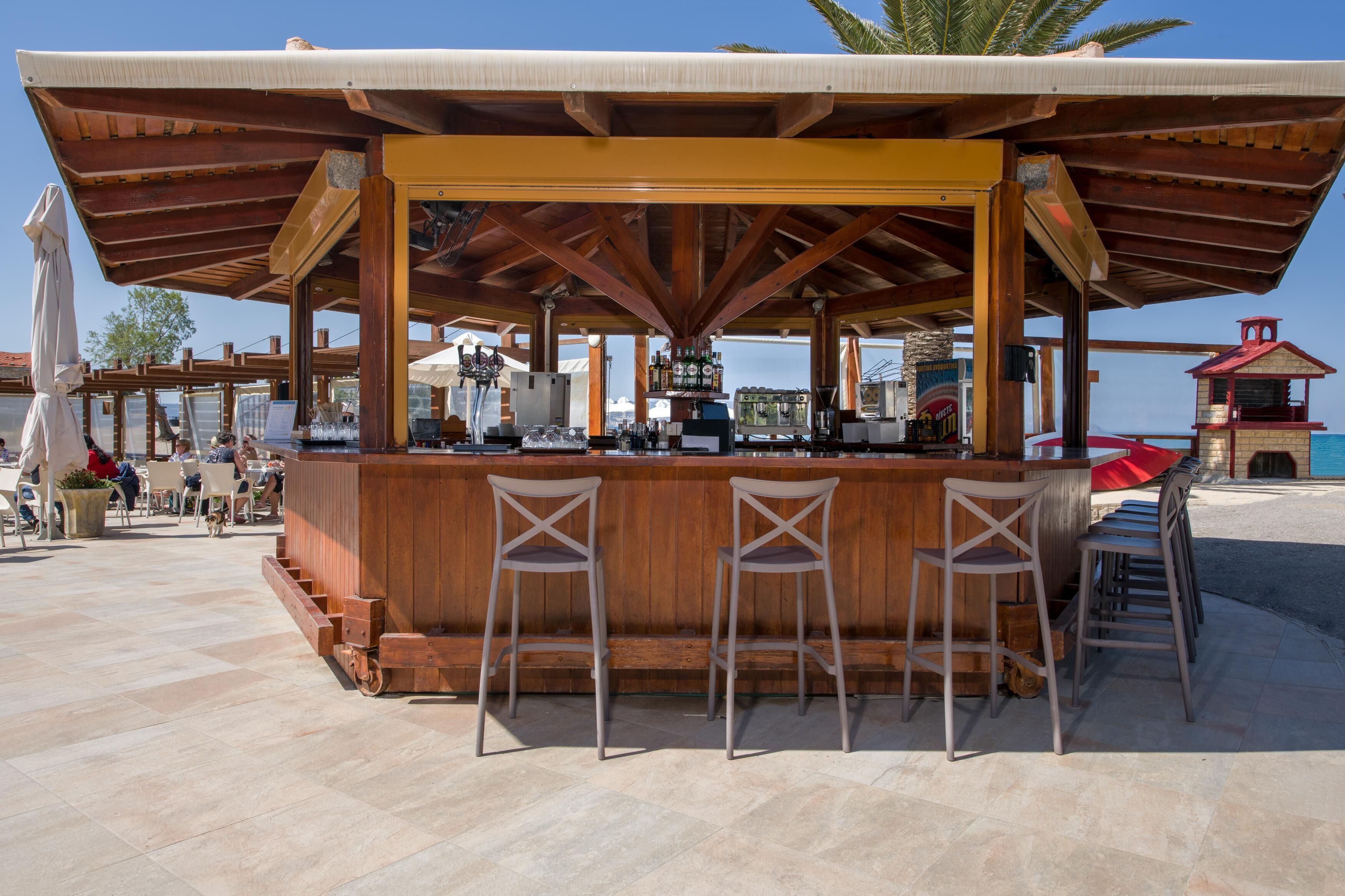Un área de bar al aire libre con taburetes de madera y una barra, rodeada por una zona de estar cubierta con vistas al mar.