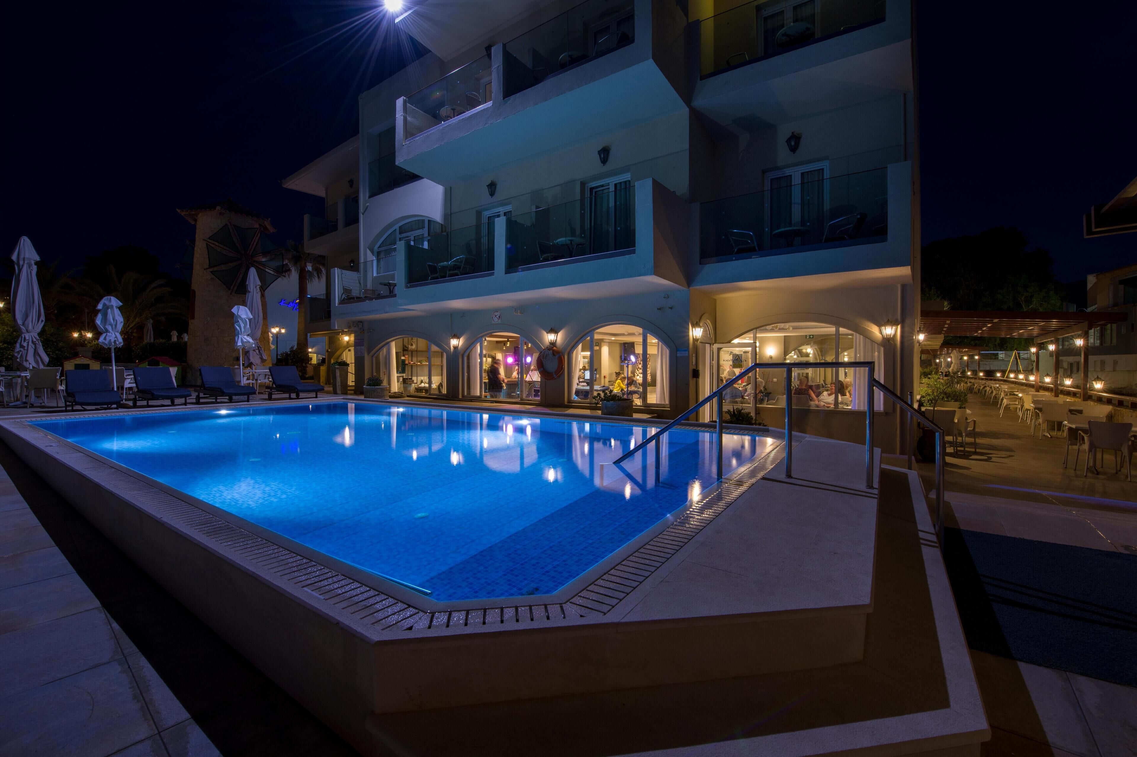 A swimming pool with lounge chairs and umbrellas, a multi-story building with balconies, and outdoor seating areas.