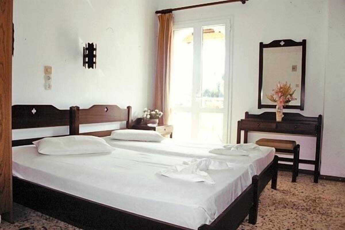 Ein Hotelzimmer mit zwei Einzelbetten, einem Holzkopfteil, einem Schreibtisch mit Stuhl, einem Spiegel und einem fenster mit Vorhängen.