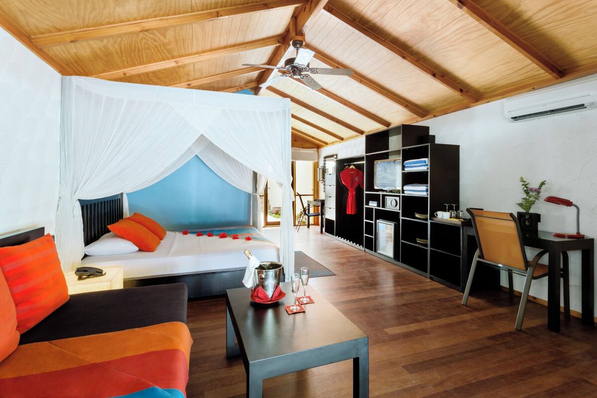 A room with a bed, a sofa, a table with a bucket and glasses, a desk with a chair, and a wardrobe.