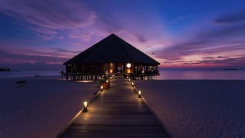 A wooden pier leading to a thatched-roof building on a beach at dusk.