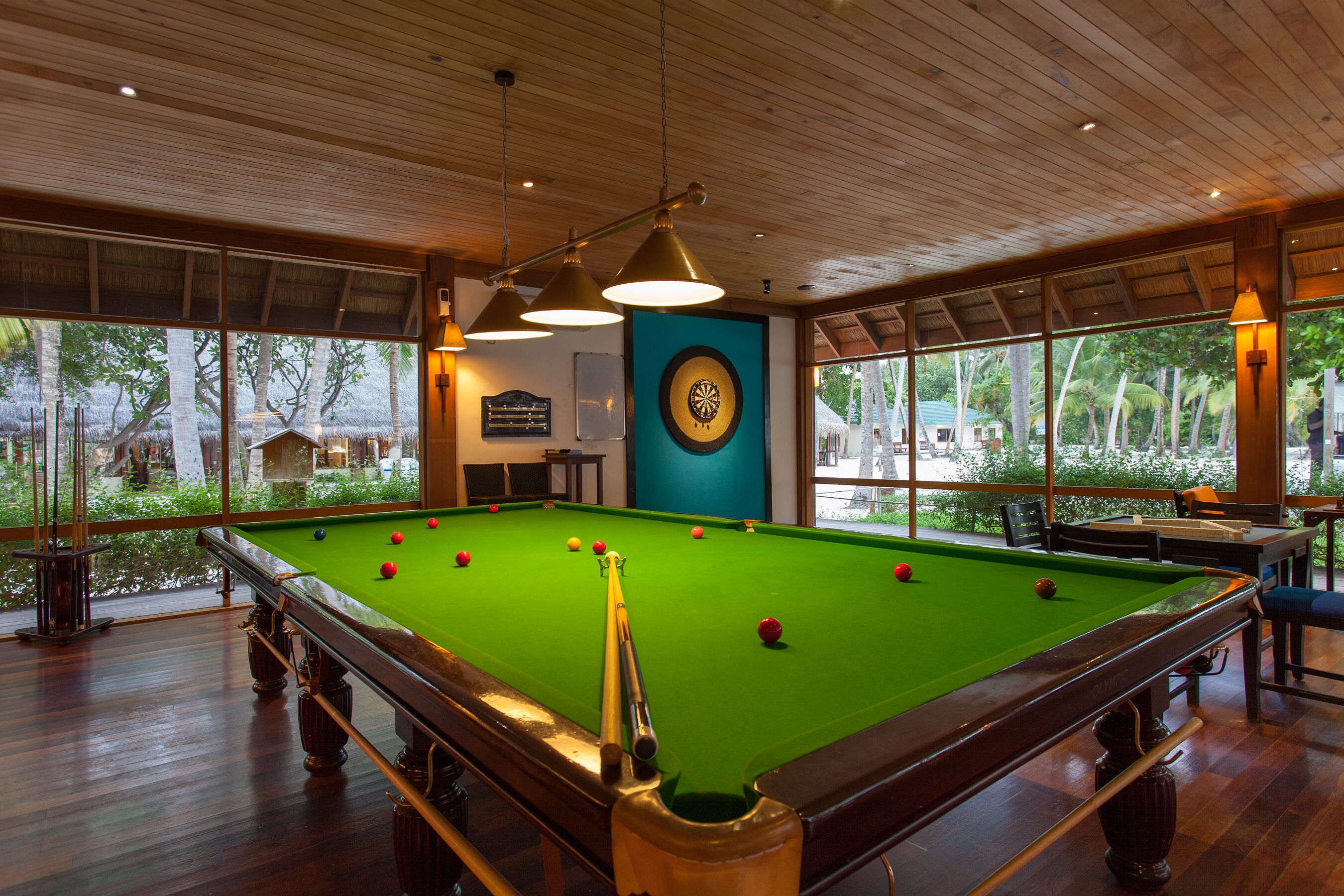 A pool table with balls, wooden ceiling, and large windows with a view of trees.