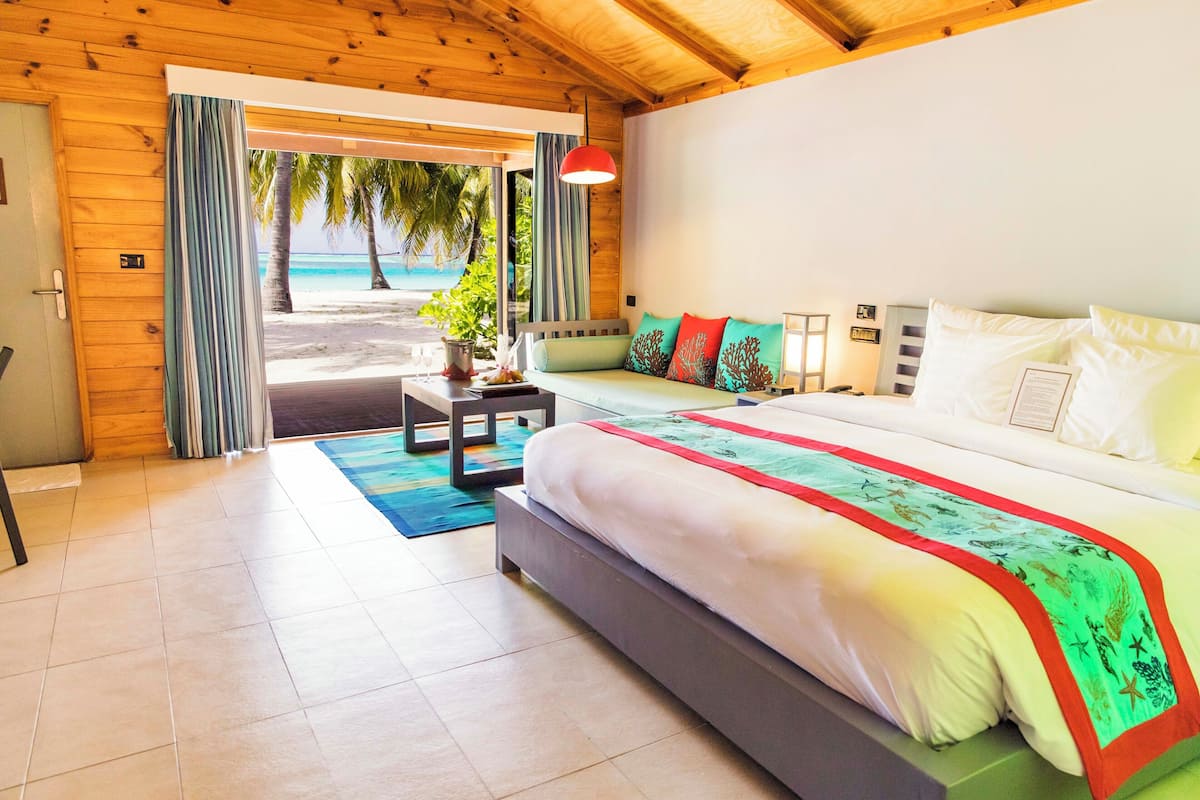 Habitación de hotel con cama, mesita de noche, lámpara y vista a la playa a través de una puerta.