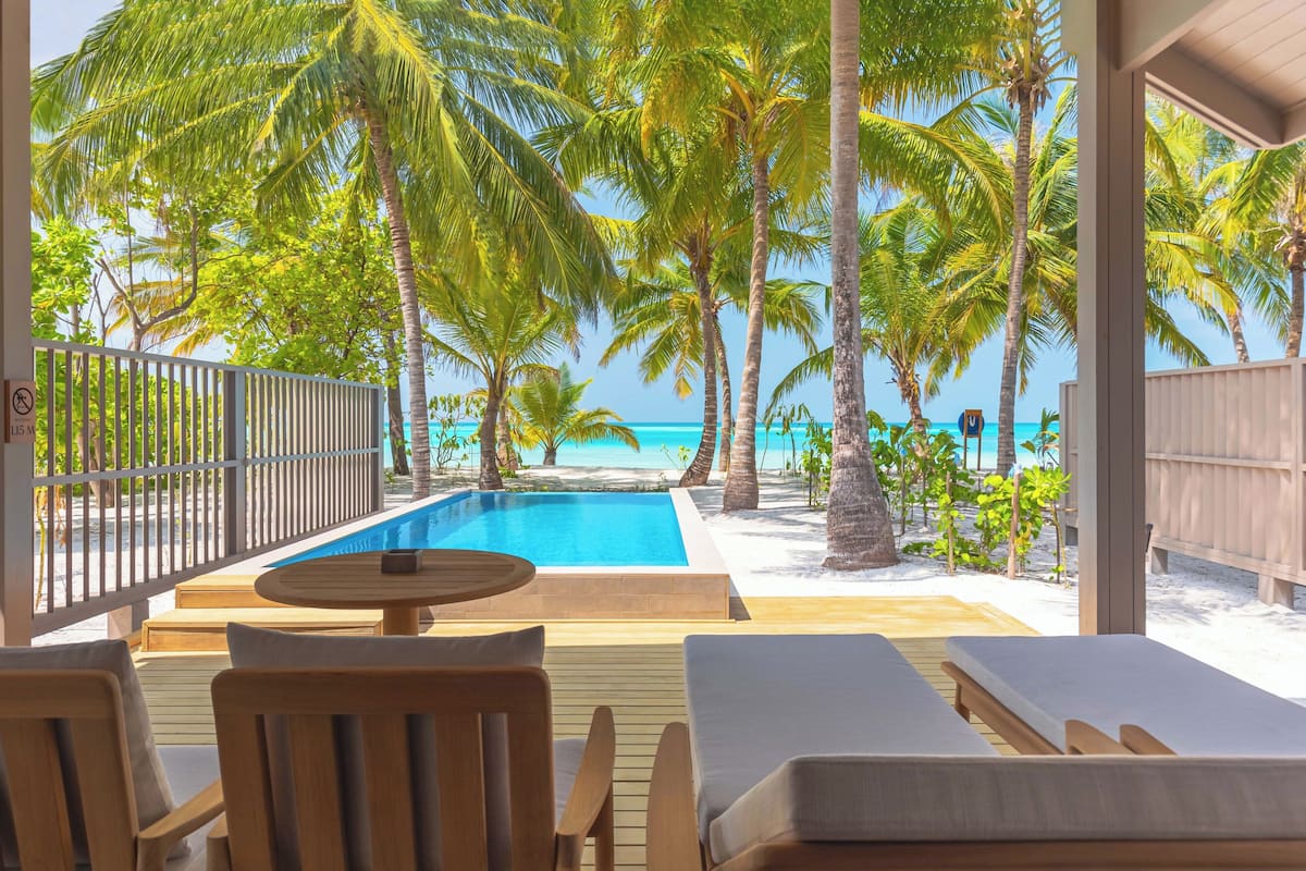 Zona de piscina con sillones reclinables y una mesa, con vistas a una playa con palmeras y un mar azul cristalino.