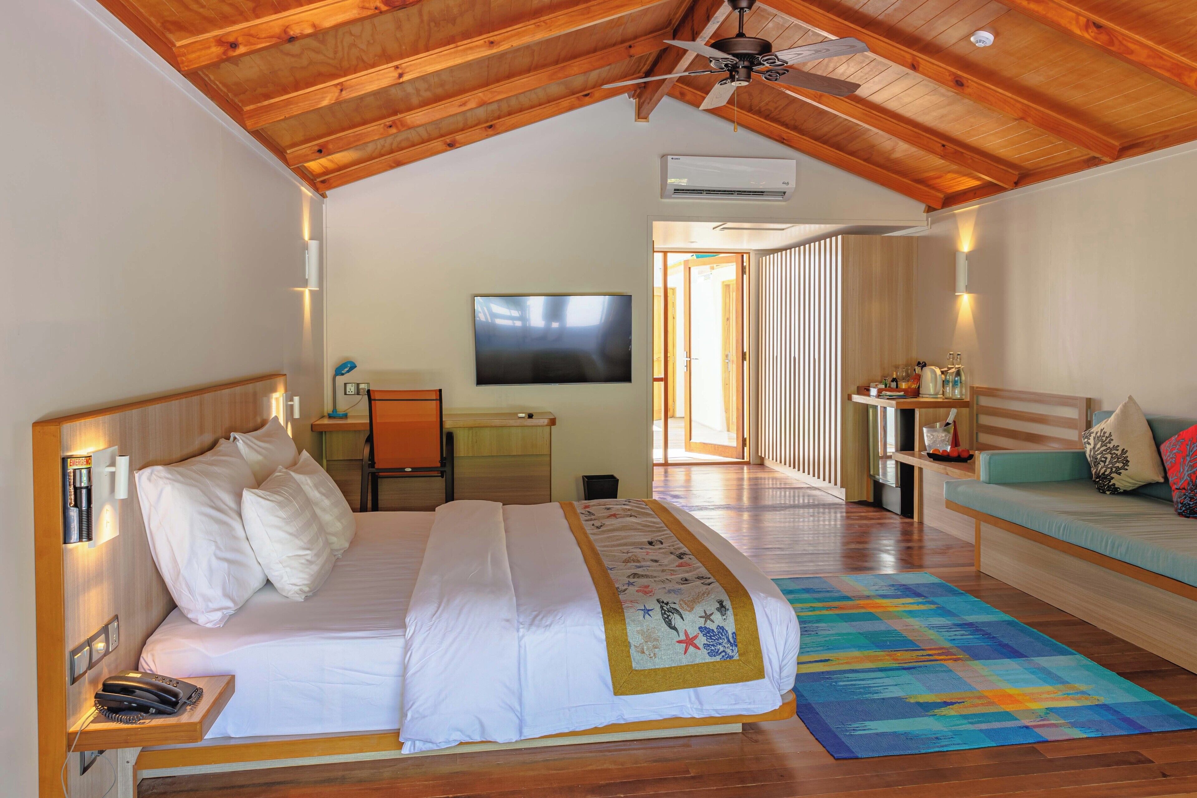 A bedroom with a bed, a TV mounted on the wall, a ceiling fan, and a wooden ceiling.