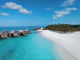 Cabañas sobre el agua conectadas por un muelle, una playa de arena blanca y un cielo azul despejado.