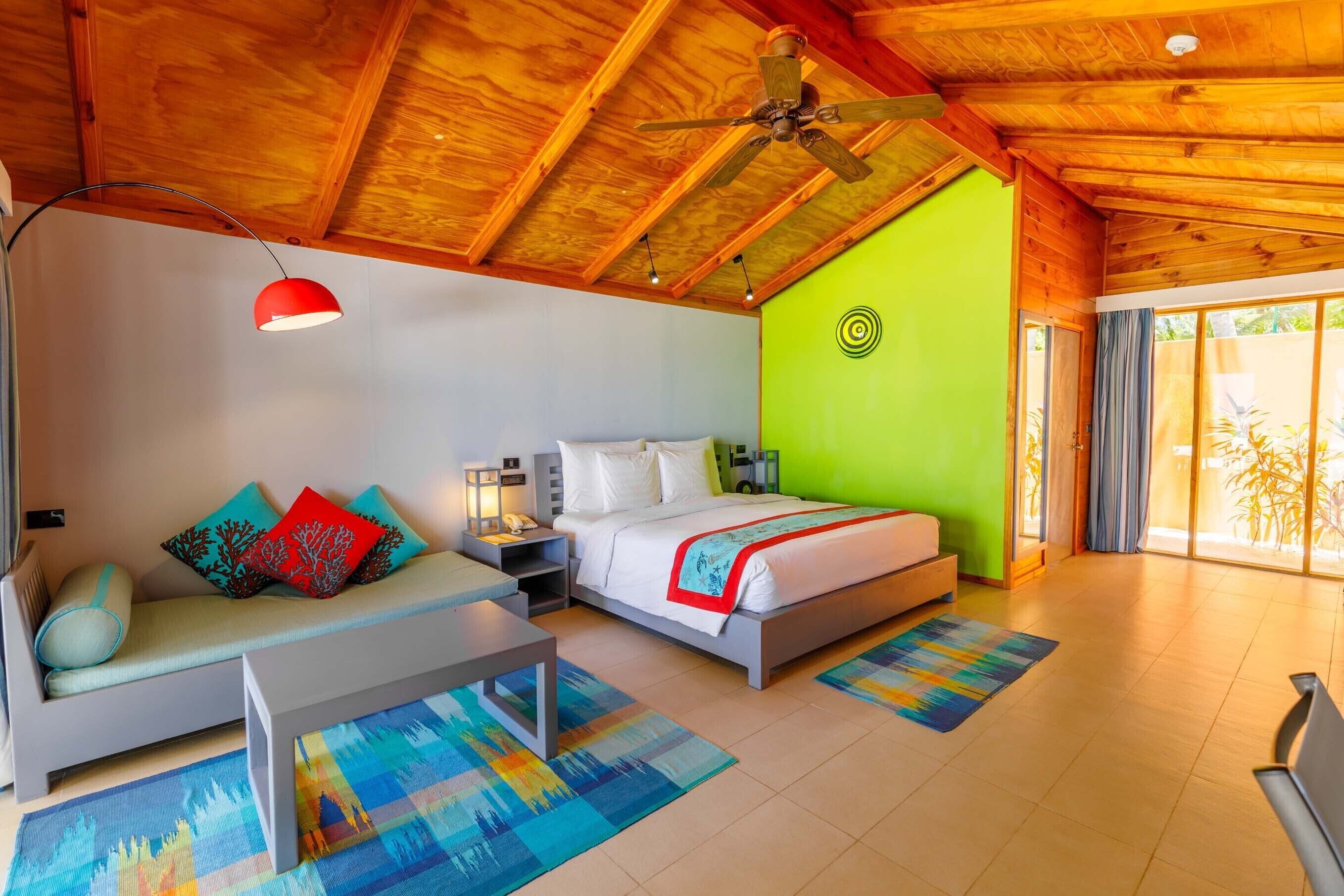 A modern hotel room with a wooden ceiling, a bed with a white and red bedspread, a grey sofa with colorful cushions, a small table, and a colorful rug.