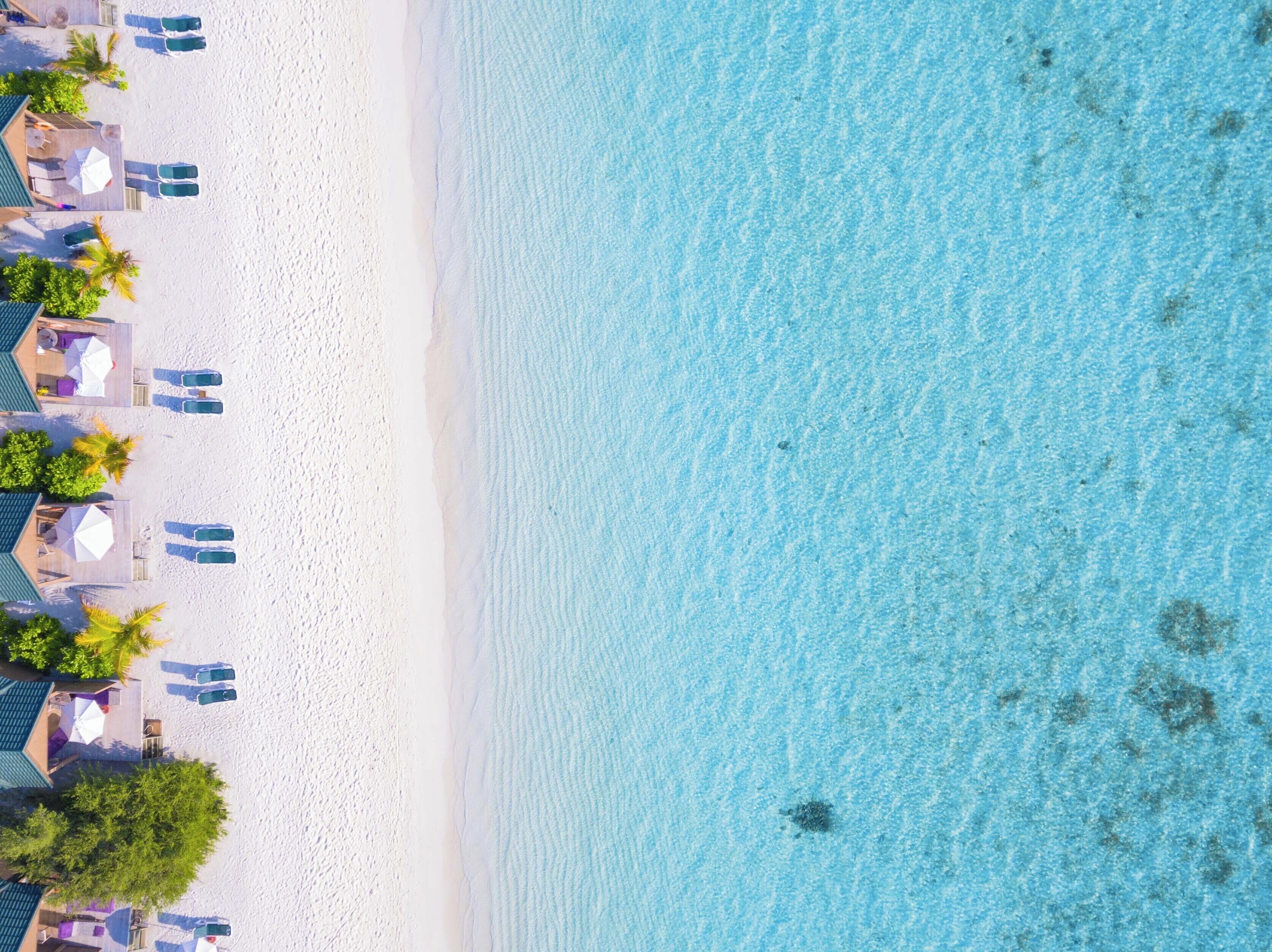 Vue aérienne d’une plage aux eaux turquoise et limpides, au sable blanc, et où l’on aperçoit plusieurs constructions le long du rivage, notamment des chaises longues et des parasols.