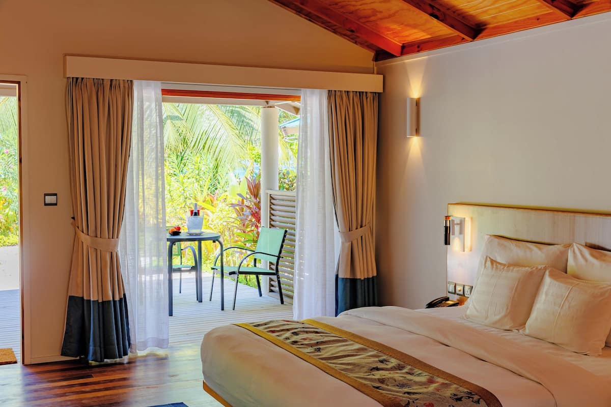 Habitación de hotel con cama, cortinas, mesita de noche y vistas a un patio con mesa y sillas.