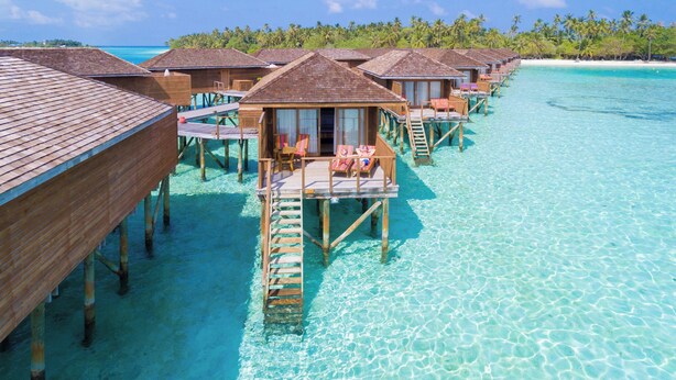 Bungalows sobre el agua con terrazas y sillones de madera, unidos por pasarelas sobre aguas turquesas cristalinas.
