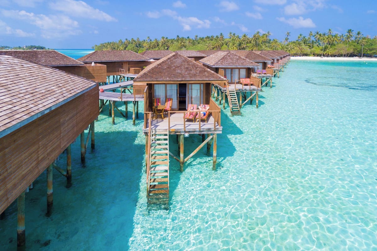 Bungalows sobre el agua con terrazas y sillones de madera, unidos por pasarelas sobre aguas turquesas cristalinas.