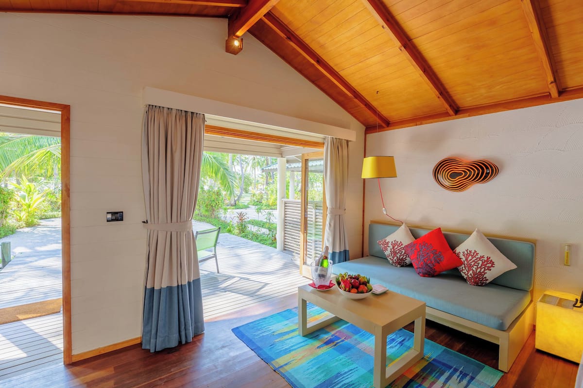 Una habitación con techo de madera, un sofá con cojines, una mesa con frutero y vistas al exterior.