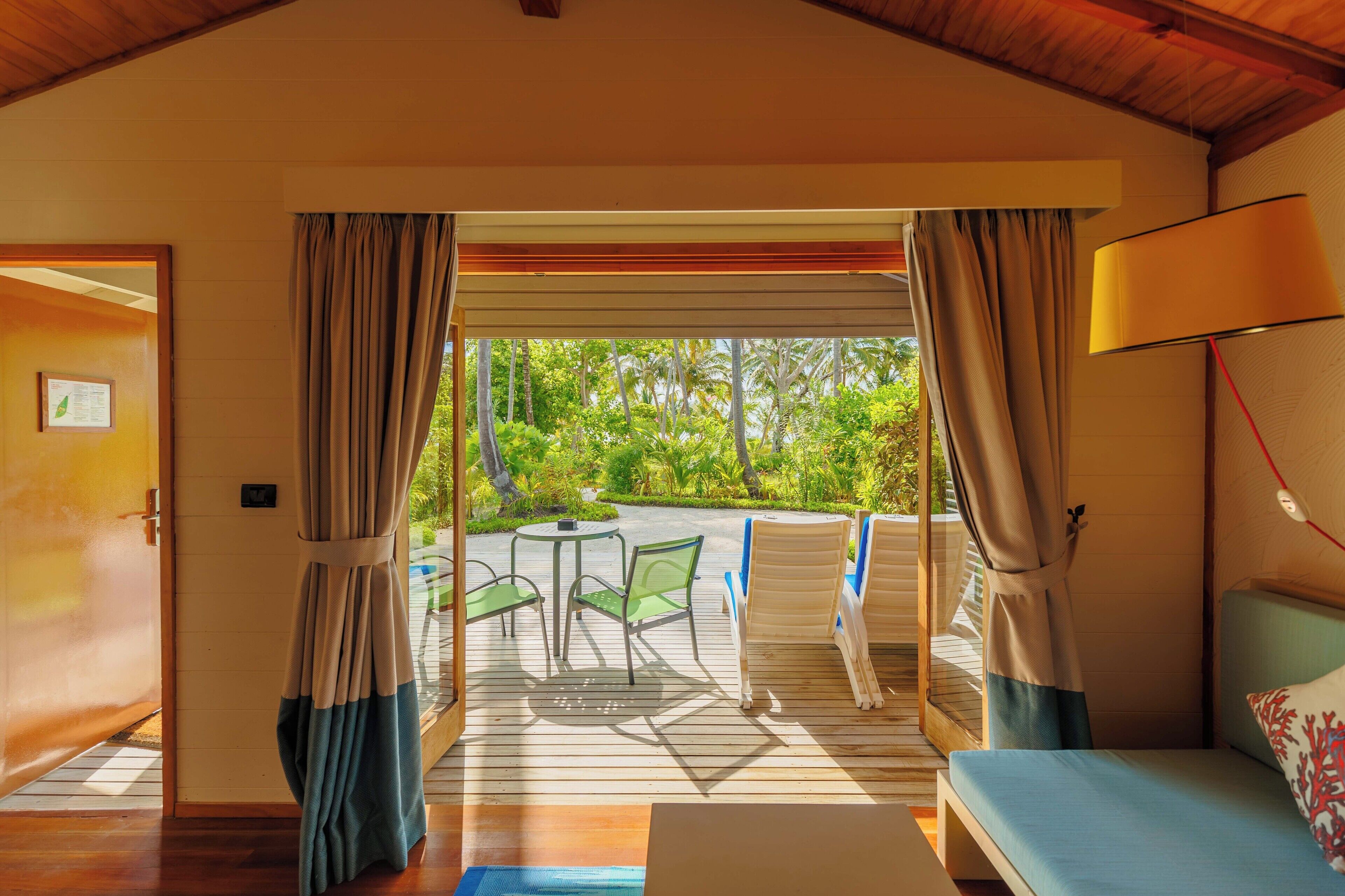 A room with a wooden ceiling, a bed with a blue mattress and a decorative pillow, a table with chairs, and a view of greenery through the curtains.